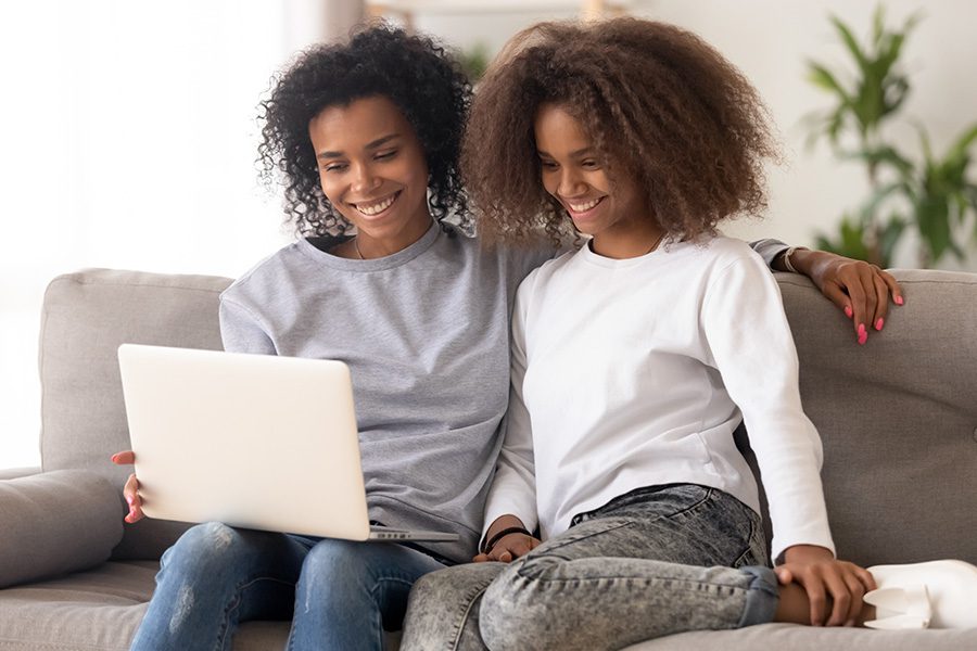 Client Center - Mother and Daughter Sitting Together and Smiling on a Sofa Using a Laptop